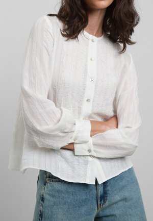 Blusa bianca a maniche lunghe con chiusura a bottoni, caratterizzata da una texture in pizzo sul davanti e tessuto a righe sulle maniche. Abbinata a jeans blu.