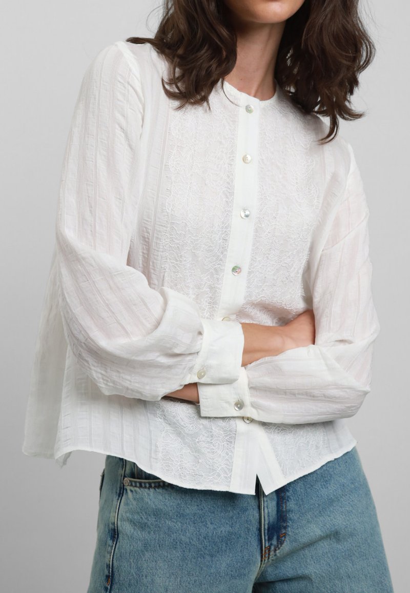 Blusa bianca a maniche lunghe con chiusura a bottoni, caratterizzata da una texture in pizzo sul davanti e tessuto a righe sulle maniche. Abbinata a jeans blu.