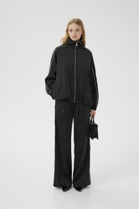Chándal de satén negro con cuello alto, que presenta rayas laterales blancas. Pantalones holgados y una chaqueta ajustada, acompañados de un bolso pequeño negro.