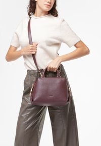 Borsa a mano in pelle color bordeaux con forma arrotondata, doppi manici e tracolla. Presenta finiture in metallo dorato e una superficie texturizzata.