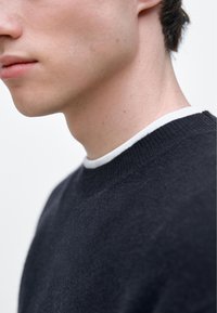 Tmavě šedý pletený svetr s bílým límcem, detailní pohled na výstřih, ukazující měkkou texturu a žebrované detaily.