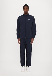 Giacca da allenamento navy con zip, logo sul petto, pantaloni abbinati e sneakers bianche. Tessuto leggero, polsini elasticizzati e design dalla vestibilità rilassata.
