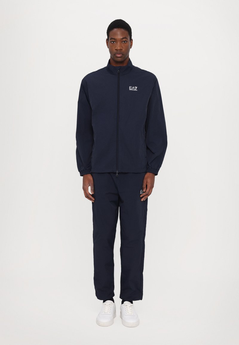 Giacca da allenamento navy con zip, logo sul petto, pantaloni abbinati e sneakers bianche. Tessuto leggero, polsini elasticizzati e design dalla vestibilità rilassata.