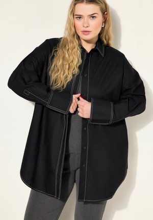 Camicia nera oversize con cuciture a contrasto bianche, fronte con bottoni, colletto a punta e maniche lunghe. Abbinata a pantaloni grigi.