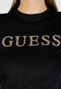 Camiseta negra de manga corta con la palabra "GUESS" en adornos de tachuelas metálicas doradas sobre el pecho. Material de algodón suave.