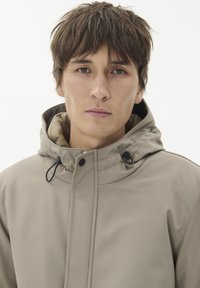 Jaunas vyras su trumpais ruda plaukais, vilkintis smėlio spalvos kaprizingą striukę su juodais raišteliais, žiūri tiesiai į priekį su neutralia išraiška.