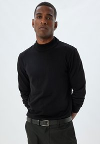 Schwarzer Rollkragenpullover aus geripptem Strickstoff, mit langen Ärmeln und einer taillierten Silhouette, getragen mit einem flachen schwarzen Gürtel.