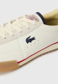 Lacoste tenisice od off-white kože s bež naglascima, sa plavim logom, crvenom prugom i gumiranim potplatom. Klasični dizajn s vezicama.