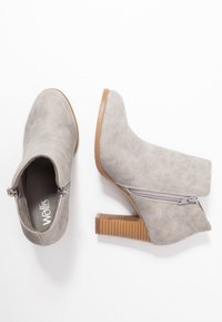 Botines de ante sintético gris con detalle de cremallera, tacón apilado y diseño de punta redondeada. La suela es de color marrón claro con textura estriada.