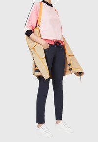 Tenue de sport comprenant un haut bicolore rose et noir, un pantalon bleu marine, et une veste beige avec des accents orange, complétée par des baskets blanches.