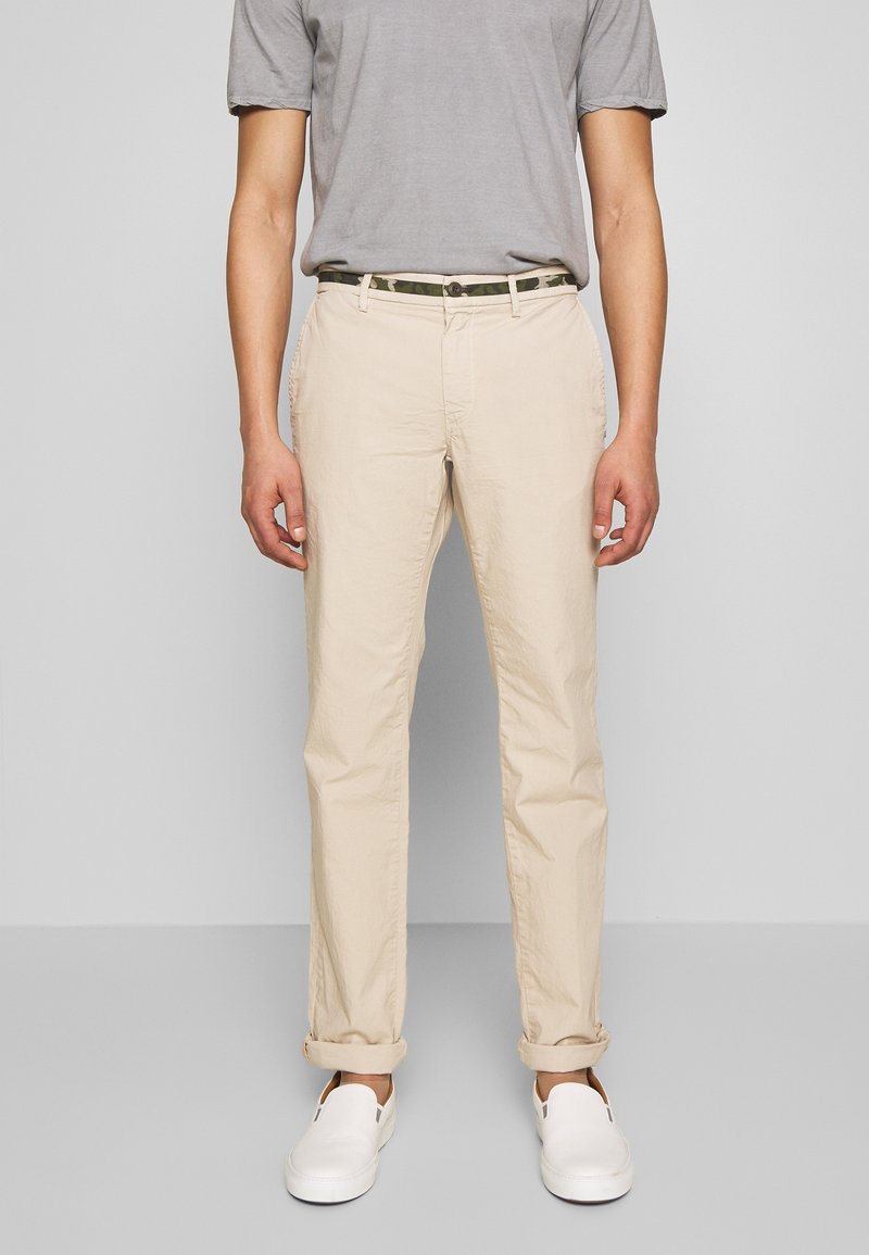 Beige cotton chinos with a straight cut, featuring front pockets and rolled cuffs, paired with a gray t-shirt and white slip-on shoes.