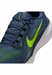Primo piano di una scarpa da corsa Nike blu con swoosh verde neon, tomaia in rete e suola bianca imbottita, che mette in mostra i dettagli dei lacci e della linguetta.