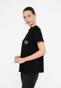 Maglietta nera a maniche corte in cotone con tasca e logo in strass. Il design è di taglio relax con scollatura rotonda.