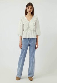Hvit bluse med volanger og V-hals, med knappelukking, paret med lyseblå vide jeans og beige sko.