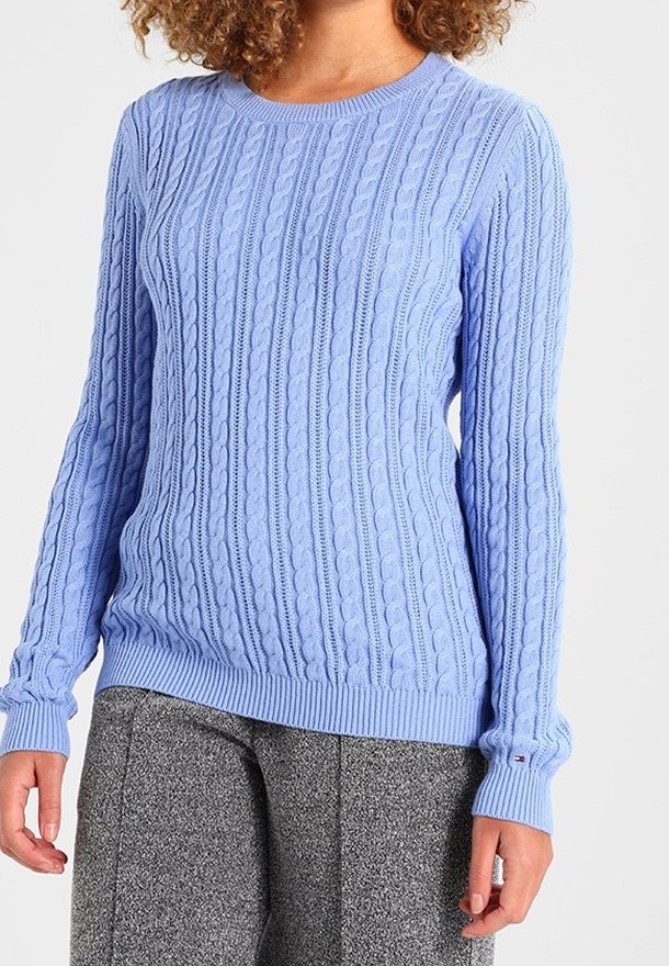 Femme portant un pull bleu clair en maille torsadée à manches longues et un pantalon gris texturé, se tenant devant un fond neutre.
