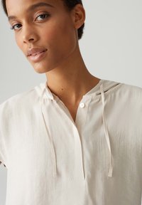 Blusa color crema con maniche corte, chiusura con bottoni e cappuccio. Tessuto morbido e texturizzato con cordini al collo.