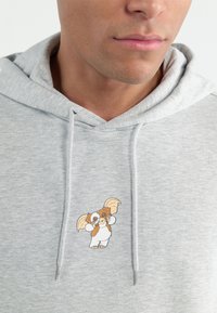 Hanorac gri din material moale, cu un mic personaj de desene animate brodat, colorat, pe piept și o buzunar frontal cu șnururi.