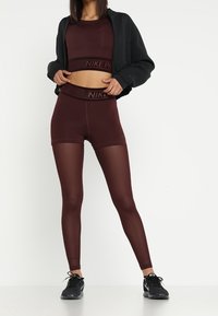 Burgunderfarbenes Sportset mit tailliertem Crop-Top und hochgeschnittenen Shorts, kombiniert mit schwarzen Turnschuhen, die mit dem Nike-Logo am Bund versehen sind.