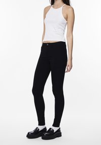 Black skinny jeans with a smooth texture, high-rise waist, and front button closure, paired with a white fitted tank top and black platform shoes.