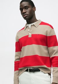 Rdeča in bež polo majica z dolgimi rokavi in ovratnikom, s tremi gumbi in majhnim logotipom; nošena s svetlimi hlačami in črnim pasom.