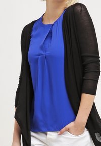 Musta neuletakki sinisen, pyöreäkauluksisen ja kevyesti laskostetun bluusin päällä. Valkoiset housut. Pehmeää kangasta, rento istuvuus.