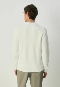 Off-white knitted sweater with a textured pattern, ribbed cuffs, and hem. Worn with light grey pants, showing a relaxed fit and crew neck.