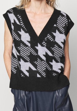 Gilet en maille sans manches noir et gris à motif, col en V profond, porté par-dessus un pantalon en cuir bleu foncé.