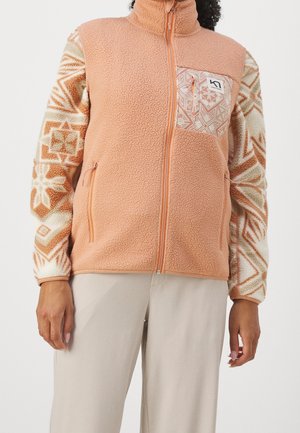 Osoba nosząca brzoskwiniową polarową kamizelkę zapinaną na zamek błyskawiczny z wzorzystą kieszenią na piersi oraz wzorzystą koszulę z długim rękawem i geometrycznymi wzorami w odcieniach brzoskwini.