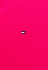 Tejido rosa brillante con un pequeño logotipo rectangular bordado en negro, blanco y rojo centrado en el marco.
