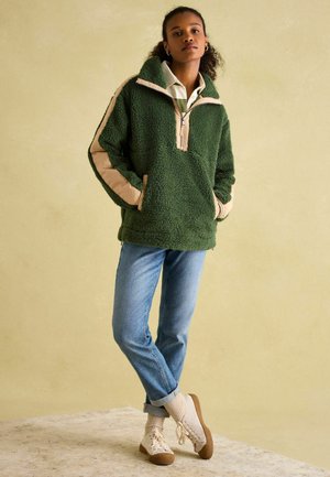 Grüner Fleece-Pullover mit Halbreißverschluss, beige Akzente an den Ärmeln. Kombiniert mit hellblauen Jeans und cremefarbenen gestrickten Schuhen.