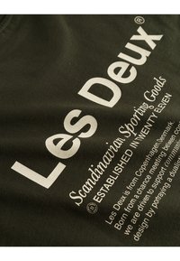 Olive green t-shirt featuring white text that includes "Les Deux" and "Scandinavian Sporting Goods," with smooth cotton texture and casual design.
