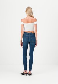 Donkerblauwe skinny jeans met een hoge taille en achterzakken, gecombineerd met een witte off-the-shoulder top. Model draagt zwarte slingback hakken.