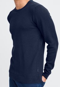 Pulover texturat de culoare bleu marin, cu guler rotund, manșete și tiv cu danteluri, realizat dintr-un material tricotat moale, cu detalii subtile ale modelului.