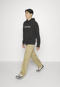 Black hoodie with "Calvin Klein" logo, khaki pants, and black and white sneakers, creating a casual outfit. Smooth fabric and relaxed fit.