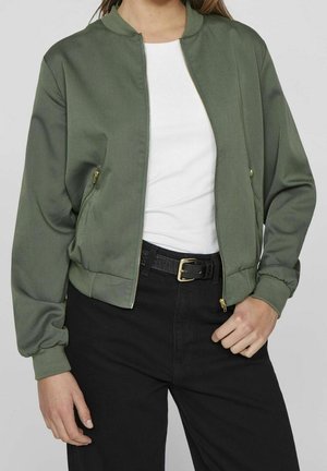 Personne portant une veste bomber verte avec une fermeture éclair dorée sur une chemise blanche, associée à un pantalon noir et une ceinture noire avec une boucle dorée.
