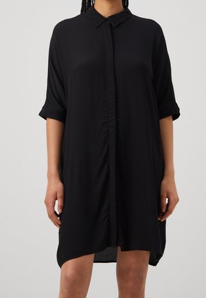 Vrouw draagt een loszittende zwart shirtjurk met kraag, korte opgerolde mouwen en een sluiting met knopen aan de voorkant.