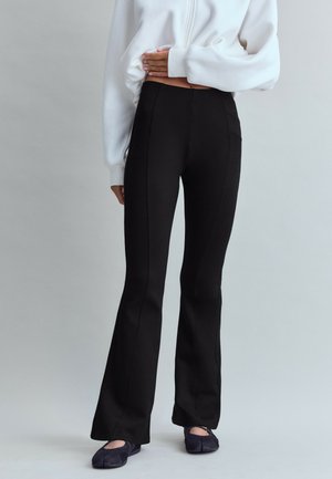 Personne portant un pantalon noir évasé, des chaussures noires sans lacets et un haut blanc court à manches longues, une main reposant sur le ventre.