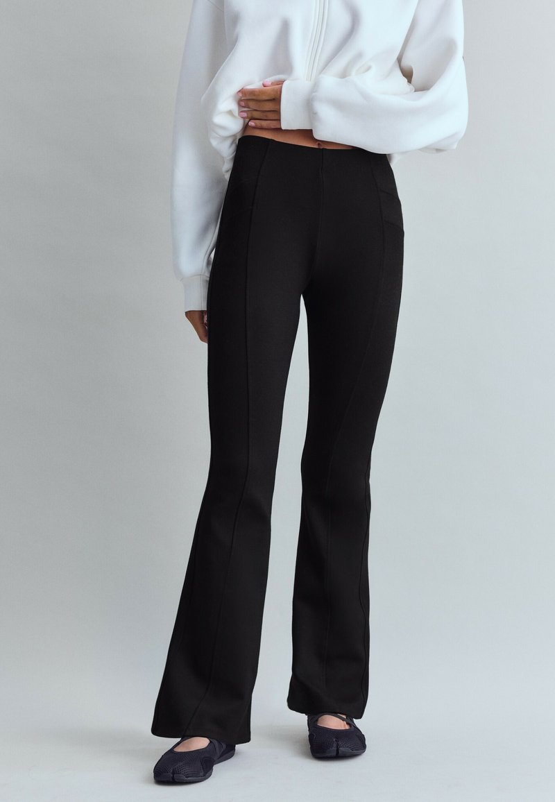 Personne portant un pantalon noir évasé, des chaussures noires sans lacets et un haut blanc court à manches longues, une main reposant sur le ventre.