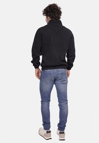 Giacca in pile nera con collare alto, polsini e vita a costine. Indossata con jeans azzurri chiari e sneakers chiare, vista dalla schiena.