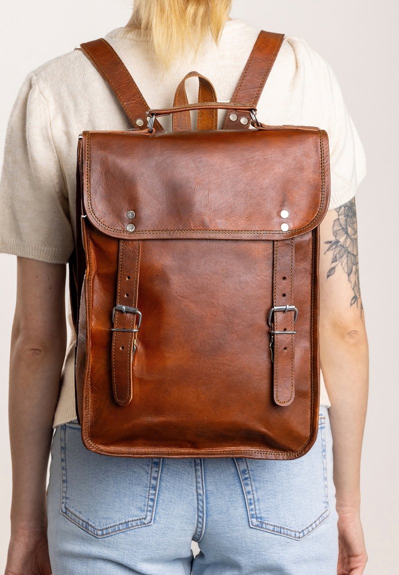 Personne portant un pull léger et un jean avec un grand sac à dos en cuir marron avec des sangles bouclées et une poignée supérieure, tatouage floral visible sur le bras.