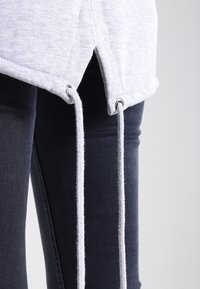 Light gray sweatshirt with split hem, featuring a drawstring threaded through silver eyelets. Paired with dark blue denim jeans.