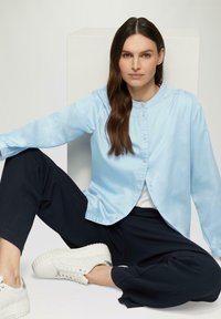 Lichtblauw, lang mouw overhemd met een ronde kraag; gecombineerd met donkere, losse broek en wit sneakers.