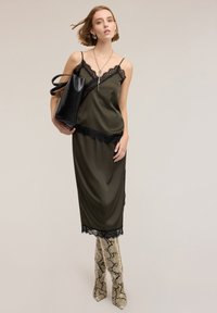 Vestito slip in satin verde oliva con dettagli in pizzo nero, abbinato a stivali al ginocchio beige con stampa serpente e una borsa nera.
