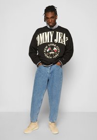 Felpa nera con logo "TOMMY JEANS", jeans in denim blu a vita alta e sneakers color crema. Presenta un colletto a girocollo e polsini a costine.