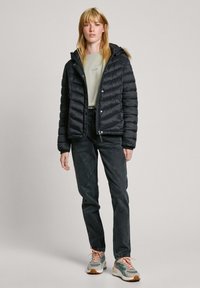 Zwarte pufferjack met kap, met horizontale stiksels, gedragen over een lichtgroen shirt en donkere spijkerbroek, gecombineerd met multikleurige sneakers.