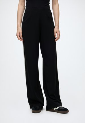 Personne vêtue d’un pantalon noir à jambes larges, d’un haut noir sans manches et de baskets noires avec des détails blancs, debout devant un fond uni.