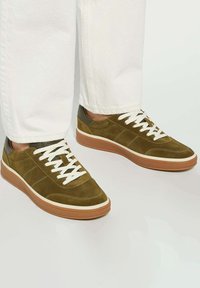 Olive green suede sneakers with white laces, light brown rubber sole, and contrasting dark accents on heel. Flat, rounded toe design.