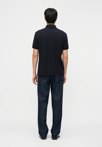 Άνδρας που στέκεται πλάτη φοράει μαύρο πόλο με κοντά μανίκια, σκουρόχρωμο μπλε τζιν και μαύρα παπούτσια σε λευκό φόντο.