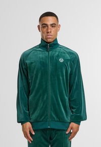 Chaqueta de chándal de terciopelo verde con cremallera, collar alto, bolsillos laterales y logo en el pecho. Textura suave y diseño de corte relajado.