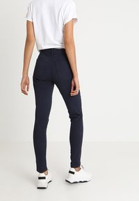 Pantalones ajustados color marino con una textura suave y cinco bolsillos. Llevado con una camiseta blanca de manga enrollada y zapatillas blancas.